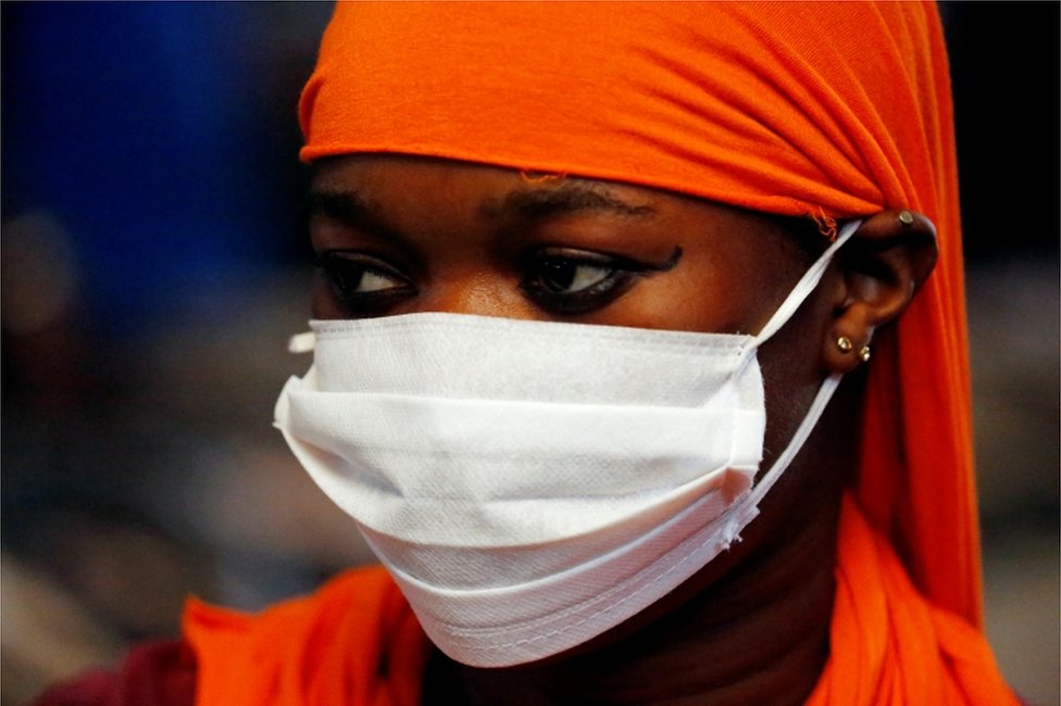A woman wears a white face mask and covers her hair with an orange fabric.
