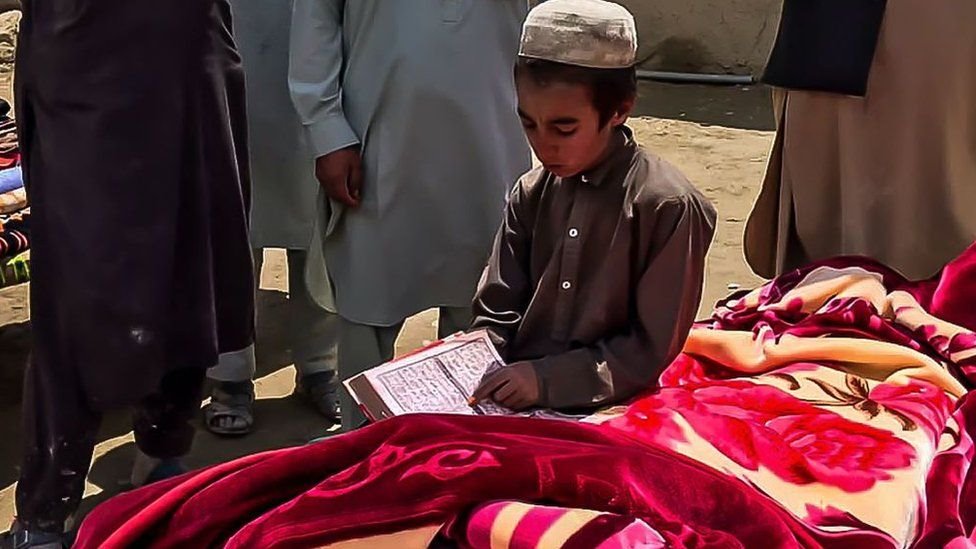 One Afghan boy dey read from di Quran while e siddon next to a body of di earthquake victim as part of burial rite