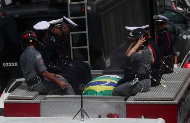 Pele coffin wey dem cover in di Brazilian flag, comot Santos stadium ontop fire engine to begin di procession as thousands of mourners line up for di streets. 