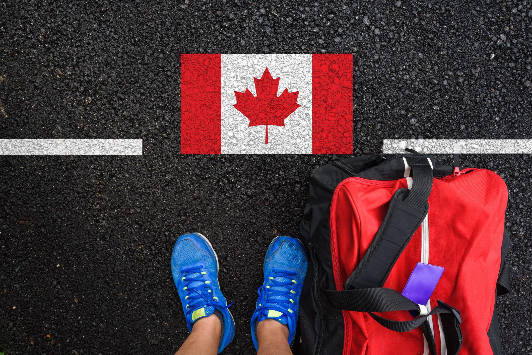 Man wit shoes and travel bag stand on asphalt next to flag of Canada and border