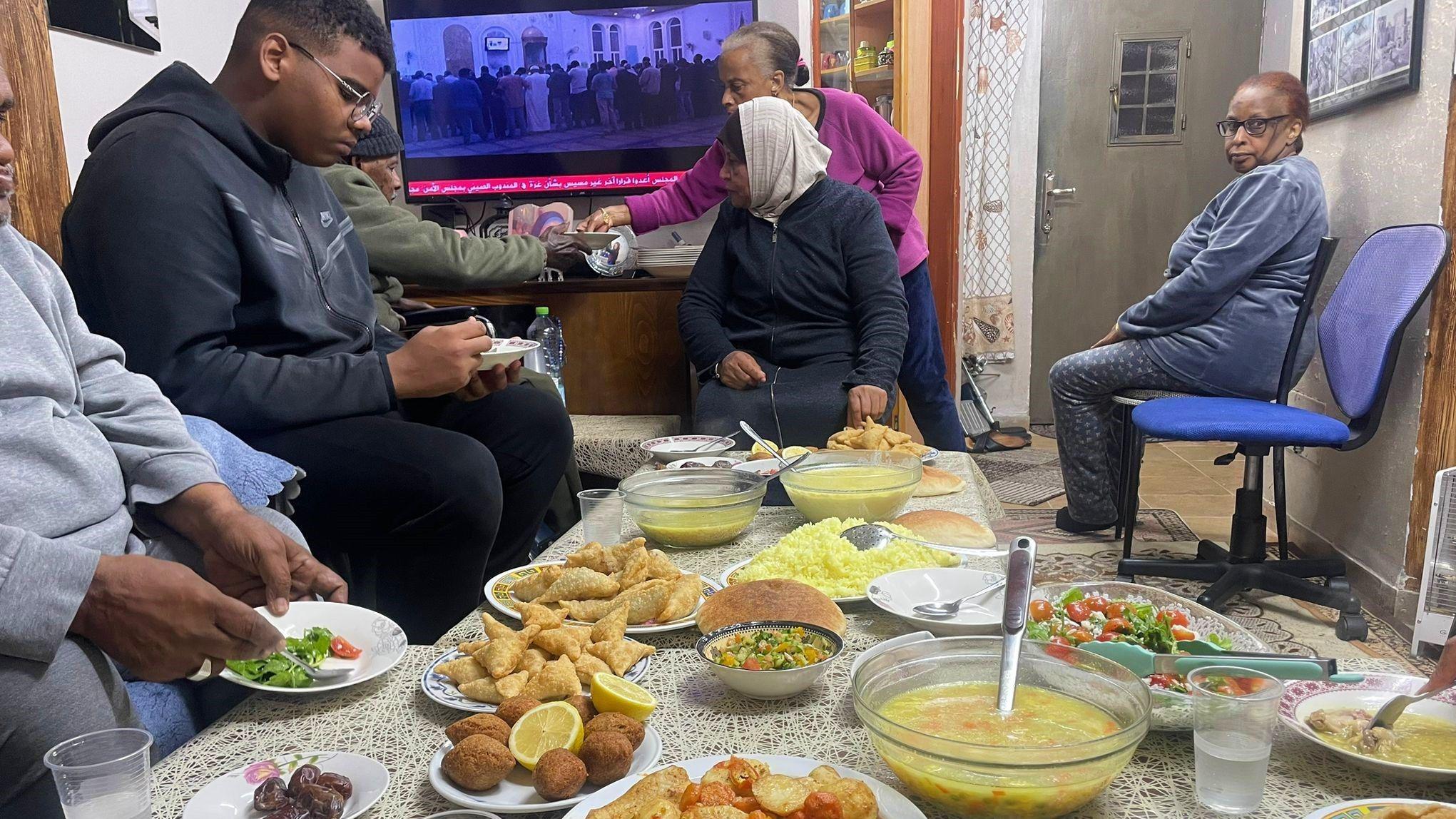 A table full of food lies at the centre of a living room with Ilham's family around it eating. 
