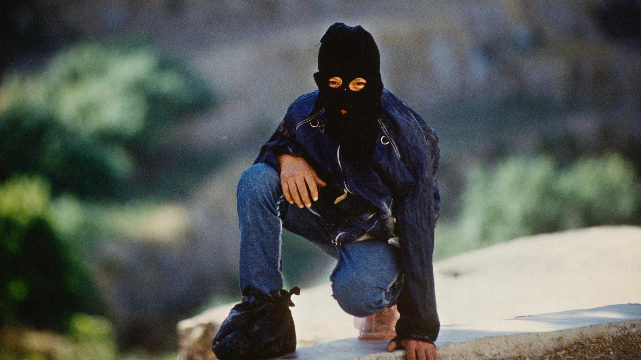 Palestinian combatants wearing a mask over his head with holes for mouth and eyes, crouching on a wall