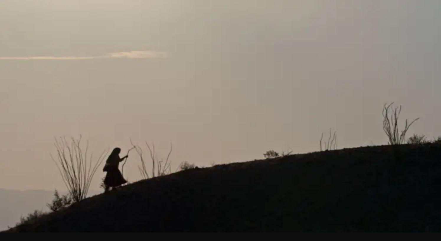 A silhouette image of a woman wearing a skirt and carrying a stick walking uphill 