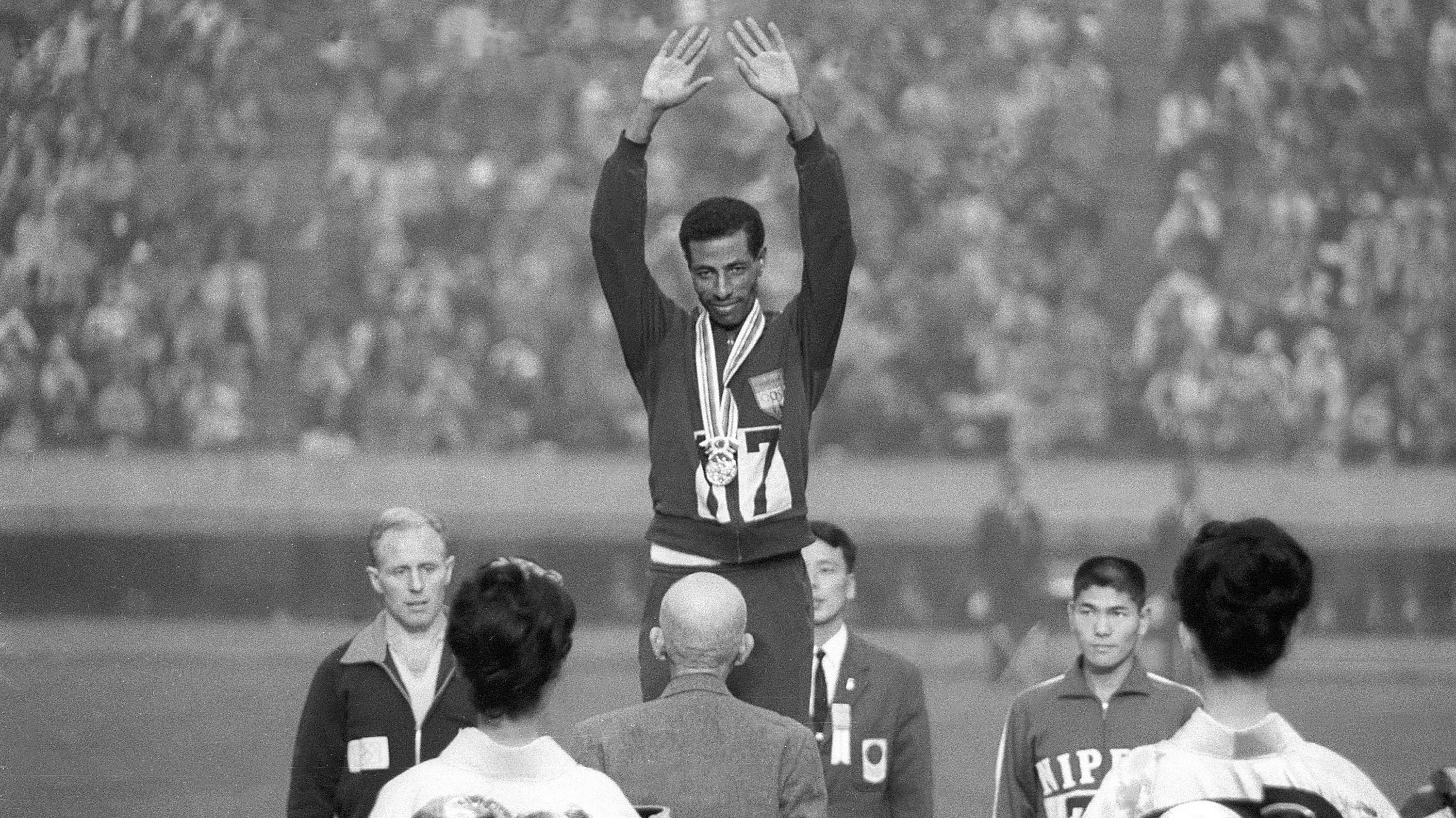 Abebe Bikila standas e raise im hands ontop podium afta e win im second Olympic gold medal