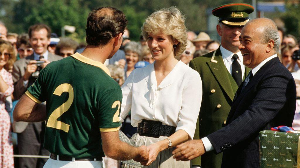 Princess Diana, for di picture dey smile for di 1980s, she dey give one glass of trophy plate to Prince Charles, wey back di camera wearing a sports t shirt wey dem write number two ontop. She get short blonde hair, and wear a white blouse and black belt. Mohamed Al Fayed dey di picture for di right of di shot, e dey smile at both of dem, for di conversation, wearing a dark suit. A crowd of spectators dey behind dem. 