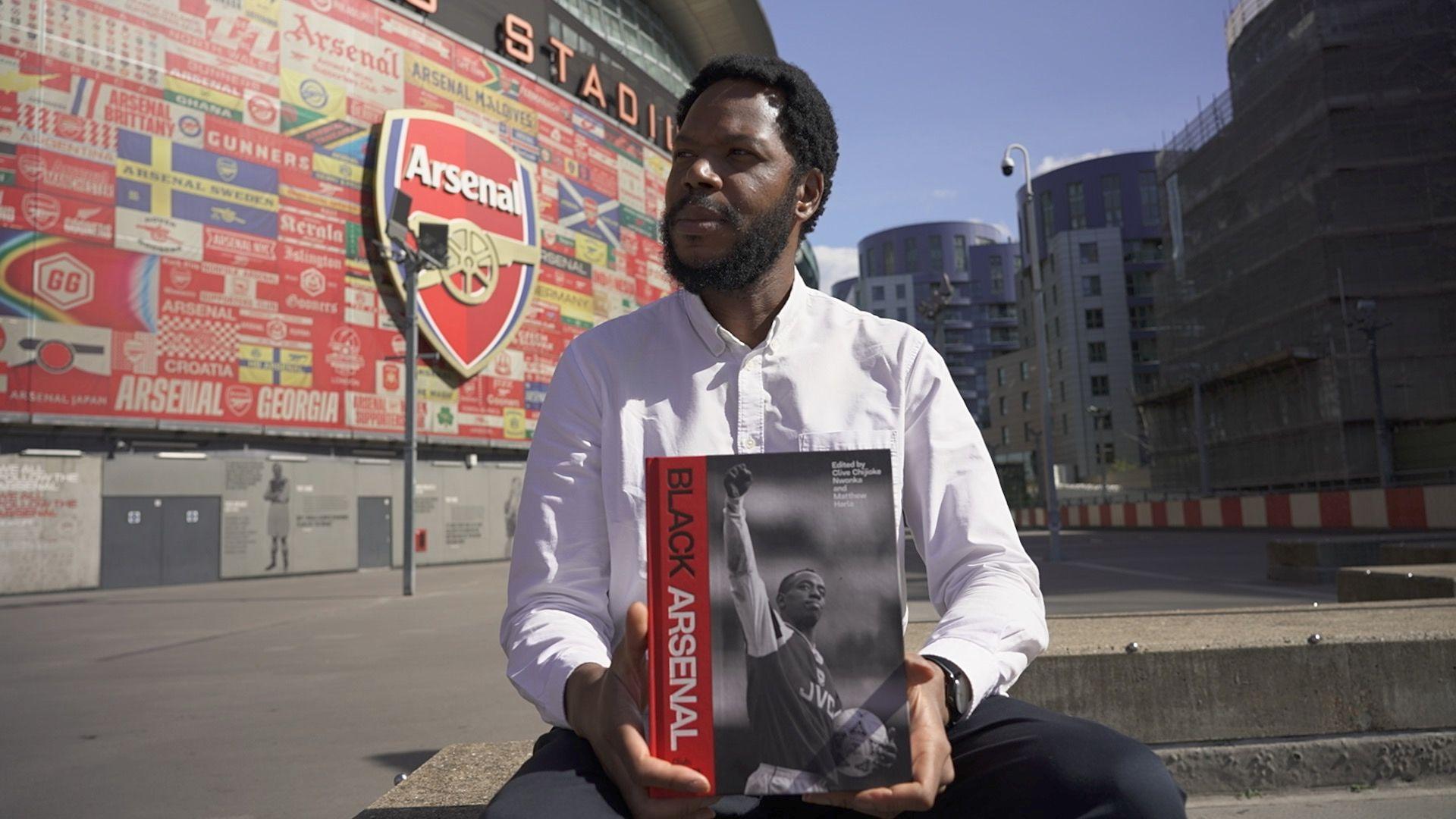 Dr Clive Chijioke Nwonka holding a copy of the book Black Arsenal outside the Emirates Stadium