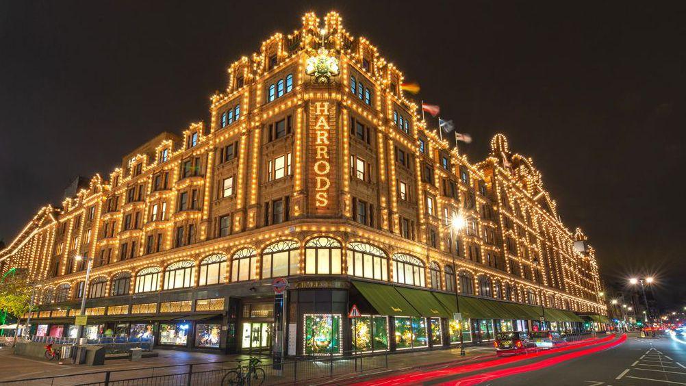 Picture of Harrods dem take in di dark. E get seven floors and many windows, with white bulbs outlining di edges. Di large shop windows on di ground floor get dark green canopy. 