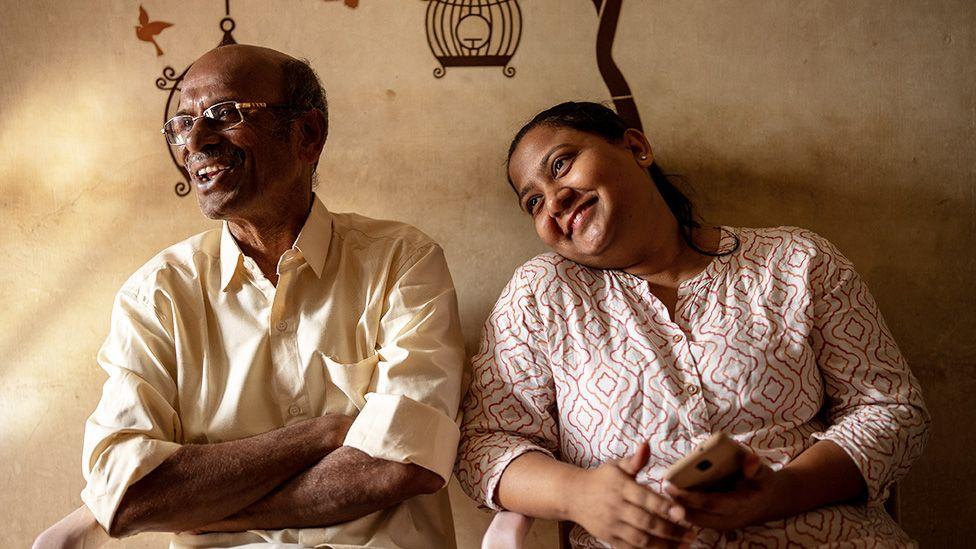 Monica for red and white blouse siddon wit her adoptive father, wey wear one cream shirt and also dey smile 