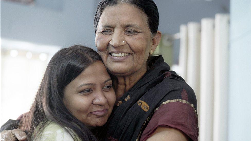 Anila - wear one black and maroon sari - and Monica hug and smile