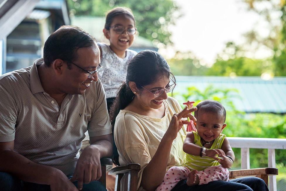 Gaurav and im wife, two of dem wear glasses and dey smile, dem hold dia baby daughter, wey wear lime green vest and pink trousers. Dia elder daughter wear black and white t-shirt, stand behind, she also dey smile 