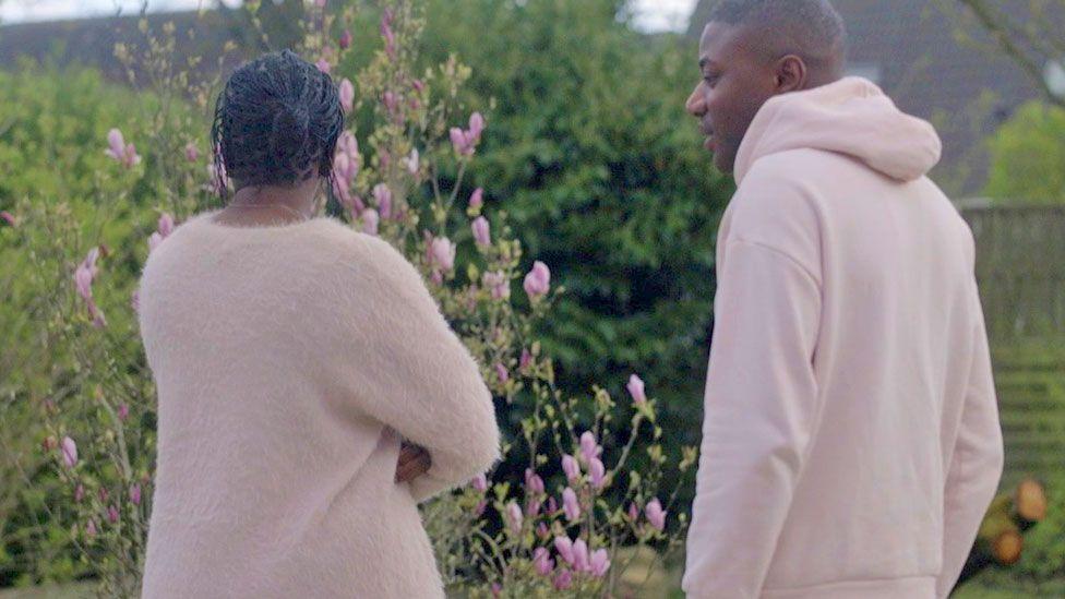 Fatoumata Sylla (l) dey look out over a flowering garden with her arms crossed and her back to di camera. Her brother Abdoul (r) dey look her - for Paris, France. Both of dem wear salmon-pink tops