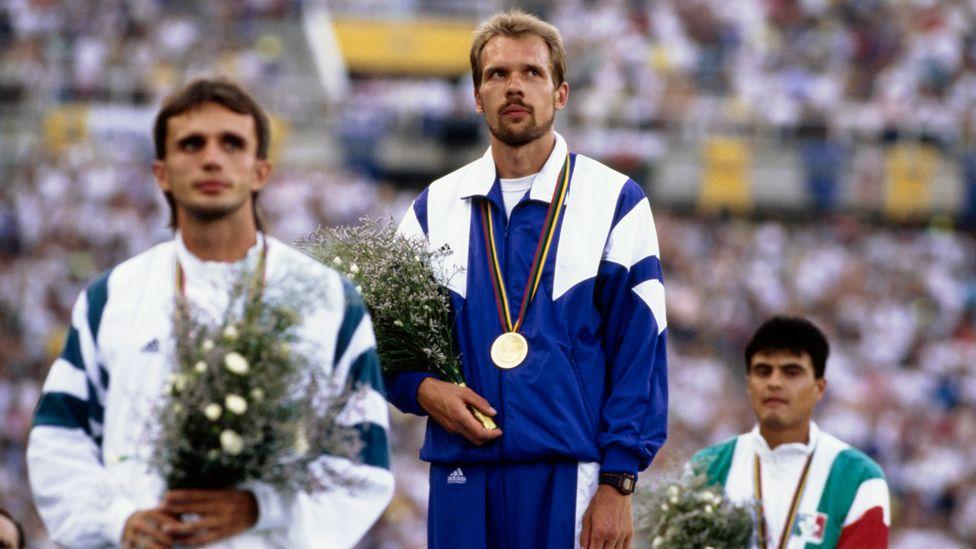 Andrey Perlov on the podium at the Olympic Stadium in Barcelona in 1992, wearing a blue and white tracksuit with a gold medal round his neck, holding a bunch of flowers -  on his left and right are the competitors who came second and third in the race