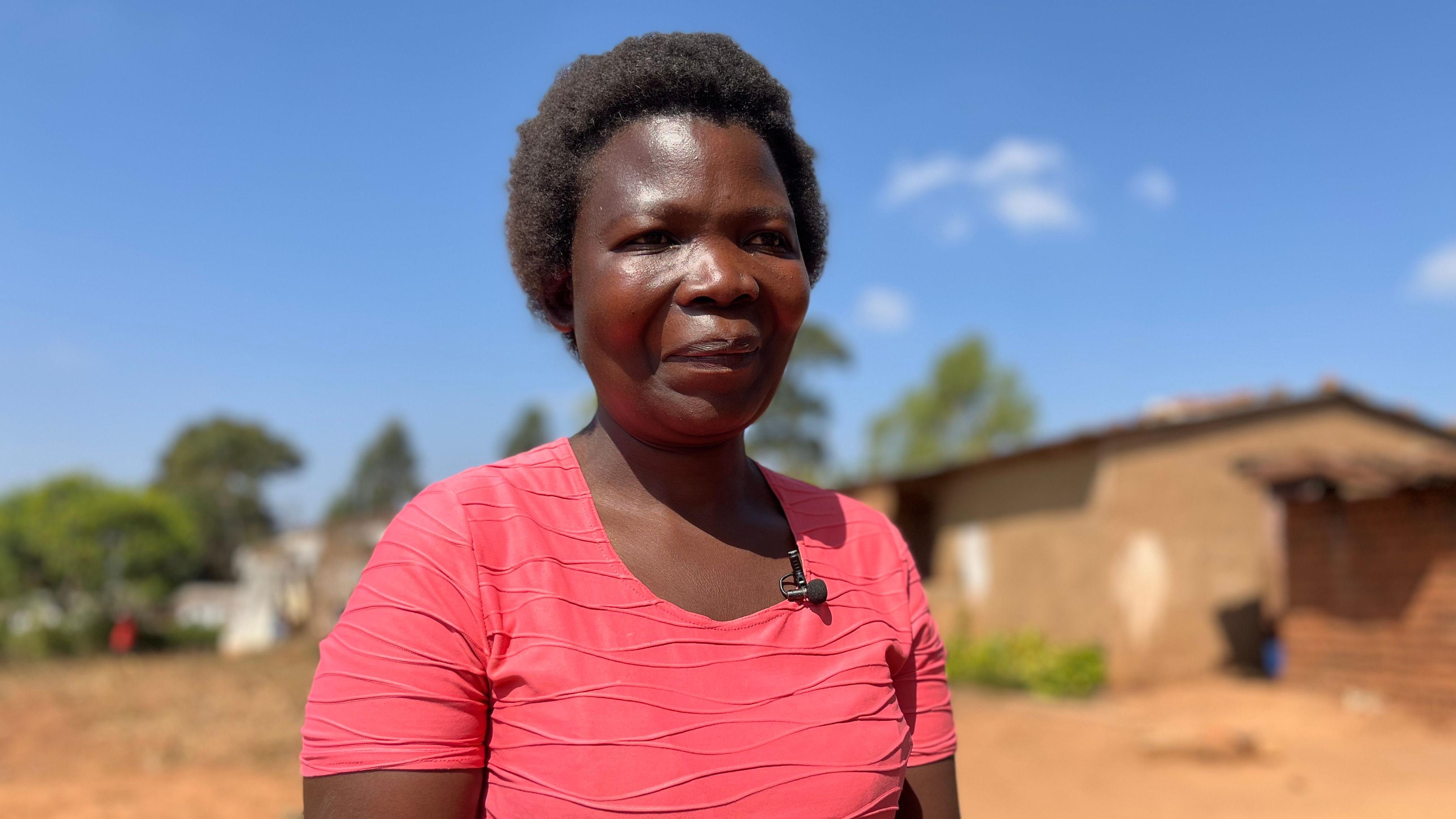 Dorothy Masasa dey stand outside, she wear pink t-shirt under di sun. Single storey building dey for background.