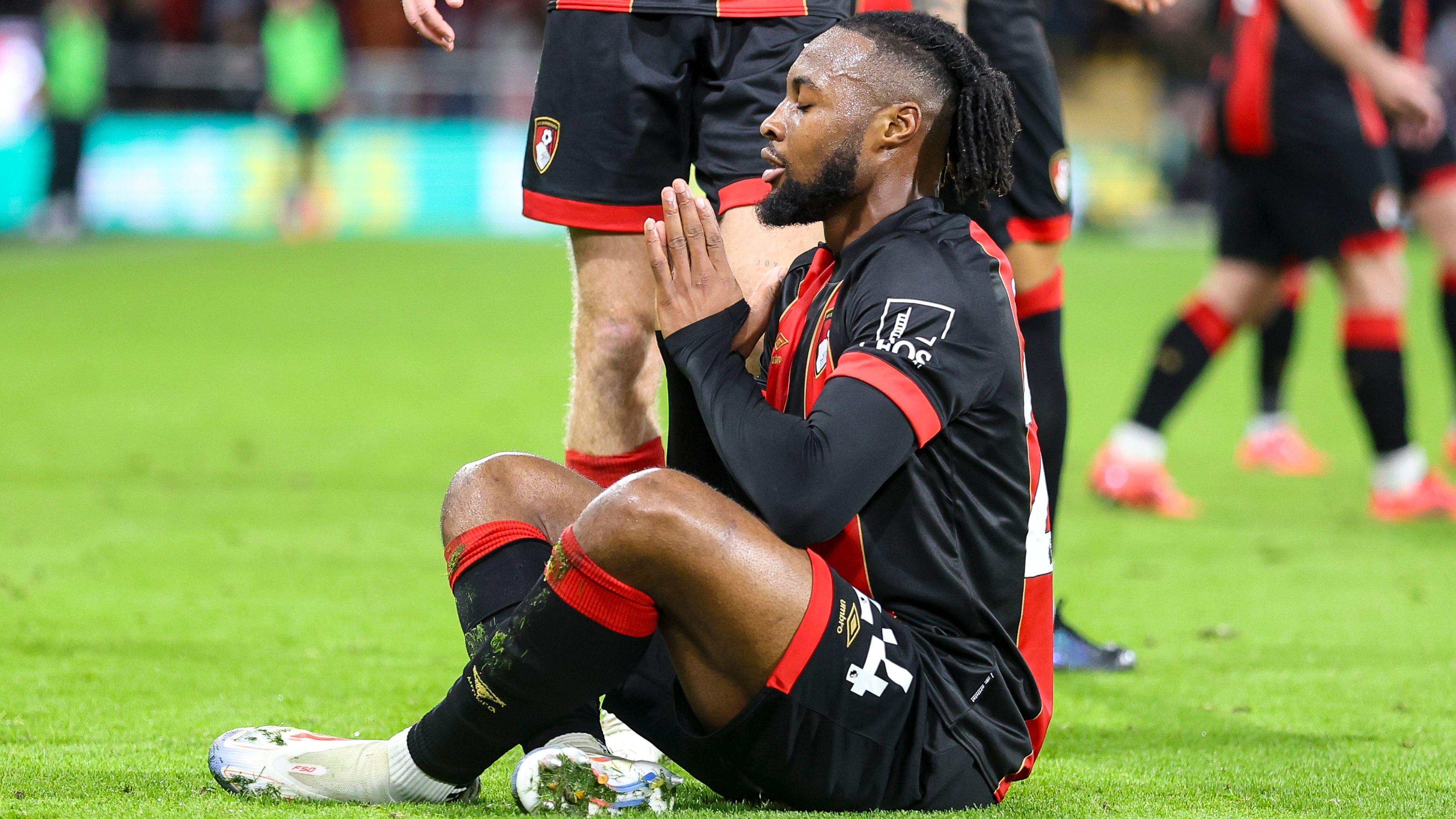 Antoine Semenyo in red and black Bournemouth kit siddon cross-leg for pitch, closes im eyes and bring im hands togeda like prayer symbol to celebrate a goal