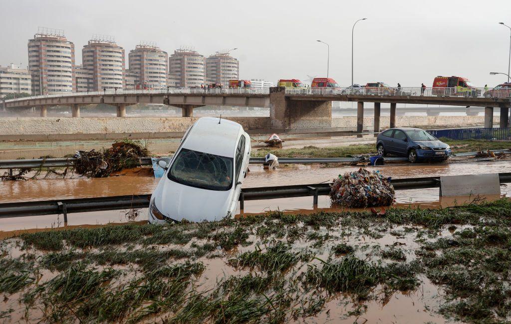 A car lies on top of a crash barrier while another car does the same on the other side of a flooded road