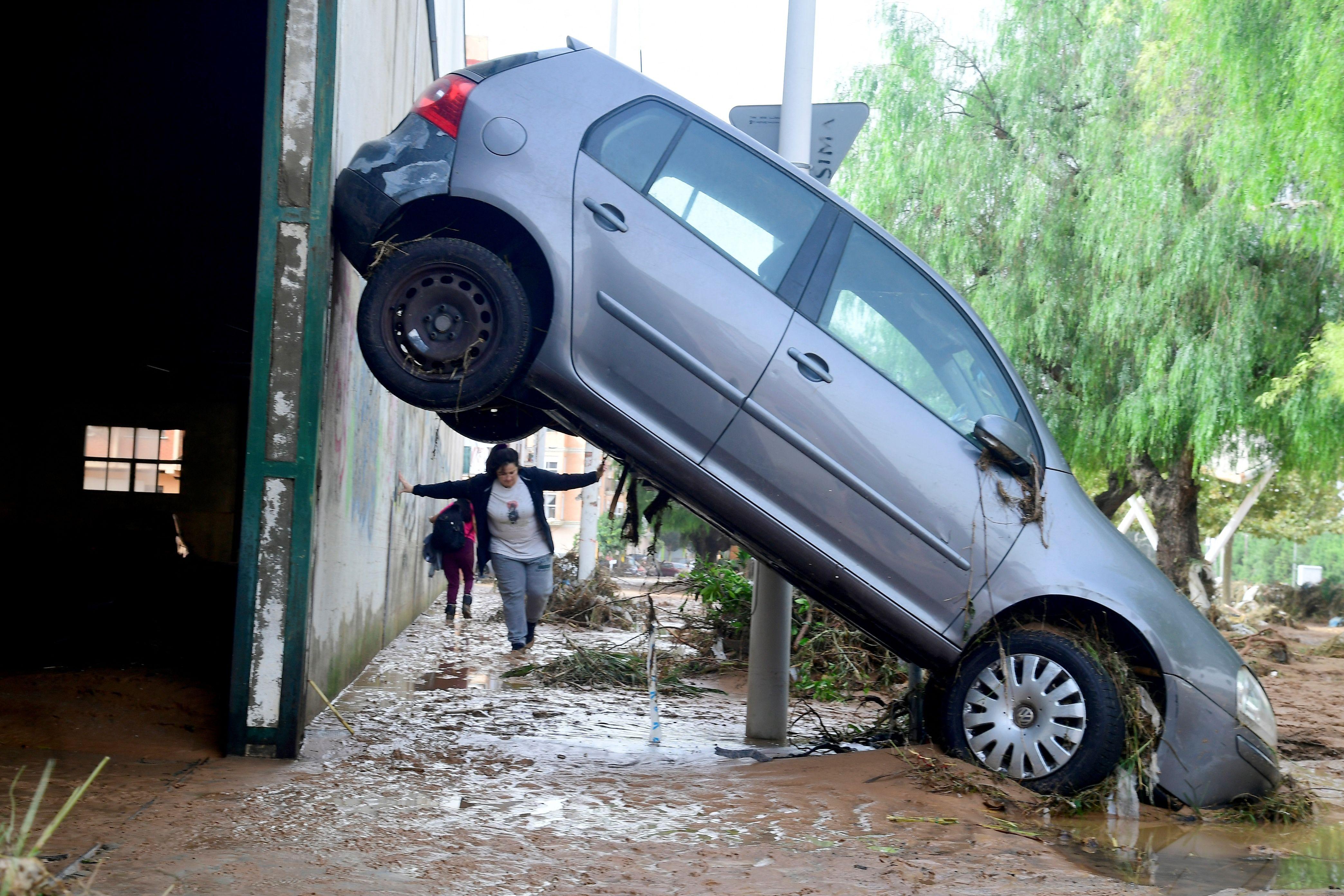 A car with its rear wheels high up against a wall and its front pointing into mud while a woman can be seen beneath its rear wheels as she walks towards the vehicle